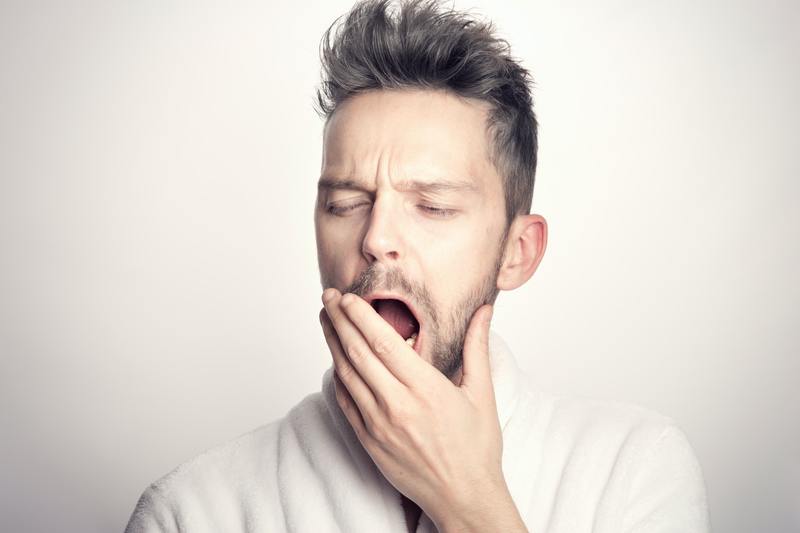 Man with brown/grey hair with a small amount of facial hair yawning and covering his mouth with his hand